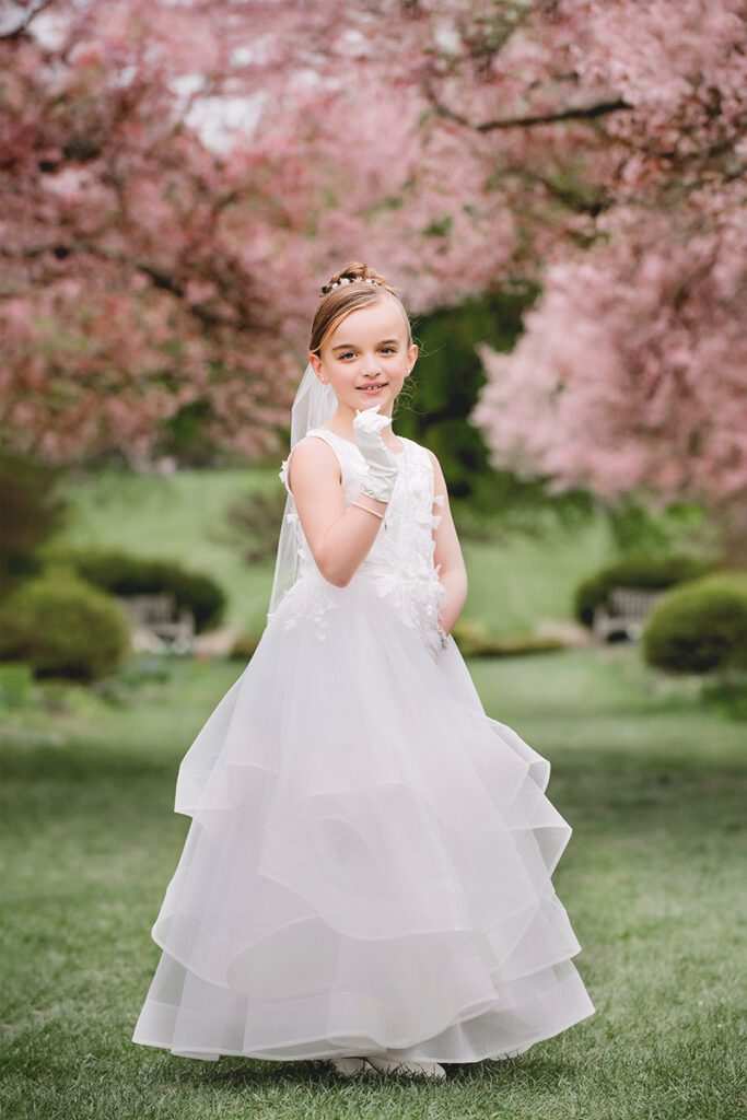First Communion photograph of a Young girl in a white dress standing in a garden with blooming pink trees, in soft natural light in New Jersey