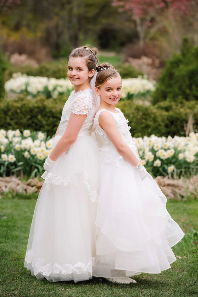 Two girls in a garden in New Jersey in First Communion dresses posing back to back in a spring with blooming flowers, and natural light.