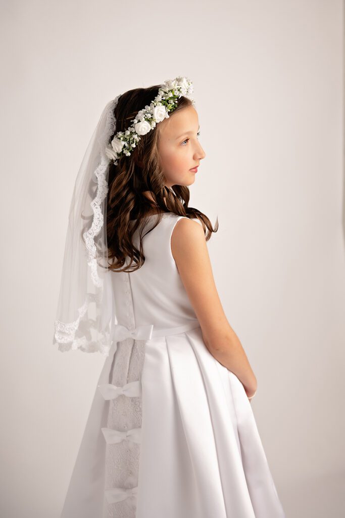 First Communion portrait of a young girl wearing a white dress, floral crown and veil, photographed in professional NJ studio with soft light.